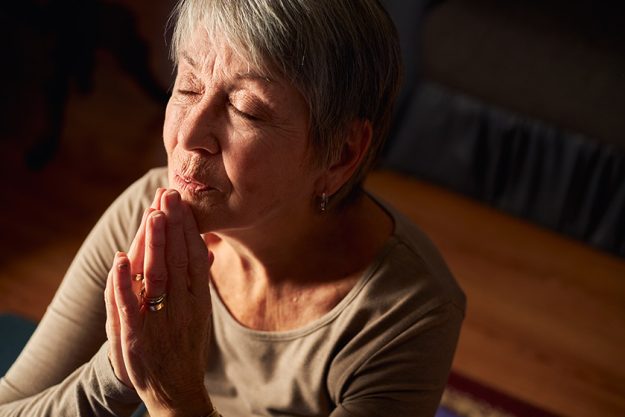 Senior Woman At Home Praying Or Meditating With Hands Together