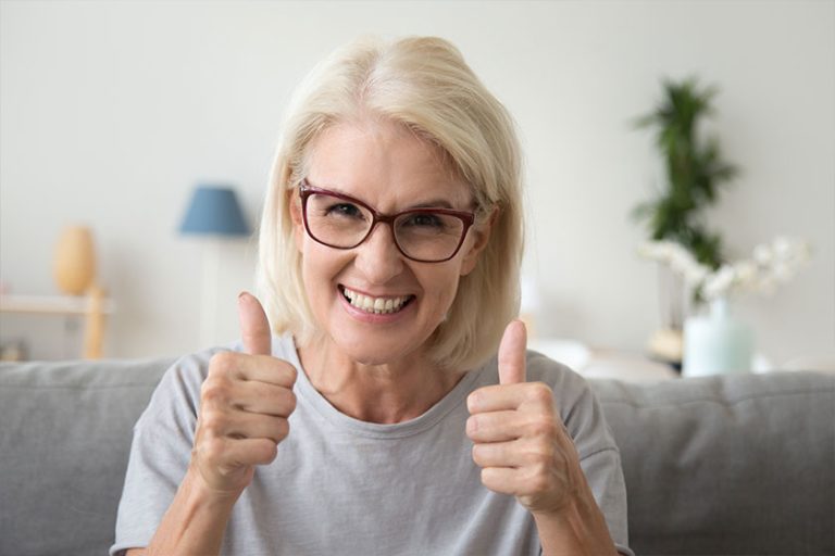 Excited mature woman in glasses sit on couch at home showing thumbs up