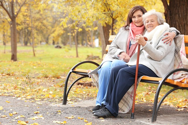 Senior woman with cane and young caregiver sitting on bench in park Senior woman with cane and young caregiver sitting on bench in park