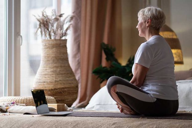 Middle age caucasian sportswoman sitting on mat with smile on face using laptop at home