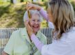 Caregiver helping senior woman to comb hair and make hairstyle when sitting on bench in park in summer.