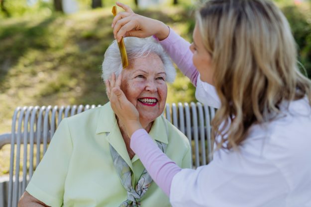 Caregiver helping senior woman to comb hair and make hairstyle when sitting on bench in park in summer. Caregiver helping senior woman to comb hair and make hairstyle when sitting on bench in park in summer.