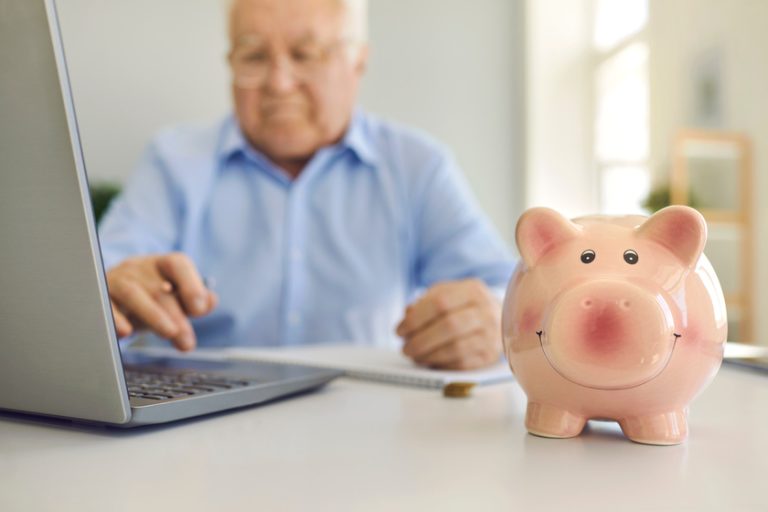 Piggy bank standing on desk with blurred senior man using laptop to pay bills online in background Piggy bank standing on desk with blurred senior man using laptop to pay bills online in background