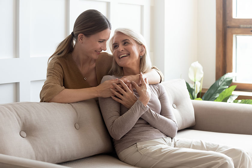 Loving adult daughter hugging older mother, standing behind couch at home