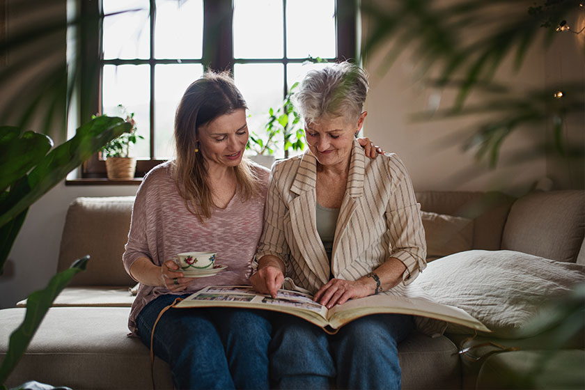 senior mother with adult daughter indoors at home, looking at family photographs