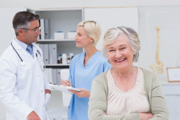Portrait of senior patient smiling while doctor and nurse discussing in background at clinic