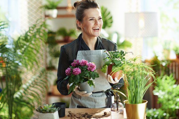 Relaxing home gardening. smiling modern 40 years old housewife in white rubber gloves with potted plant in the modern living room in sunny day.