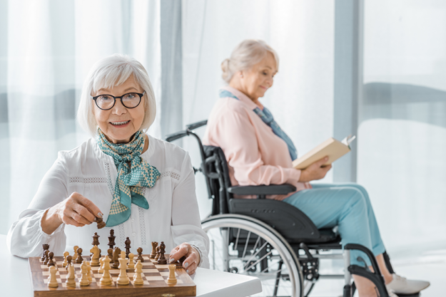 Senior woman playing chess at table while another siting wheelchair reading
