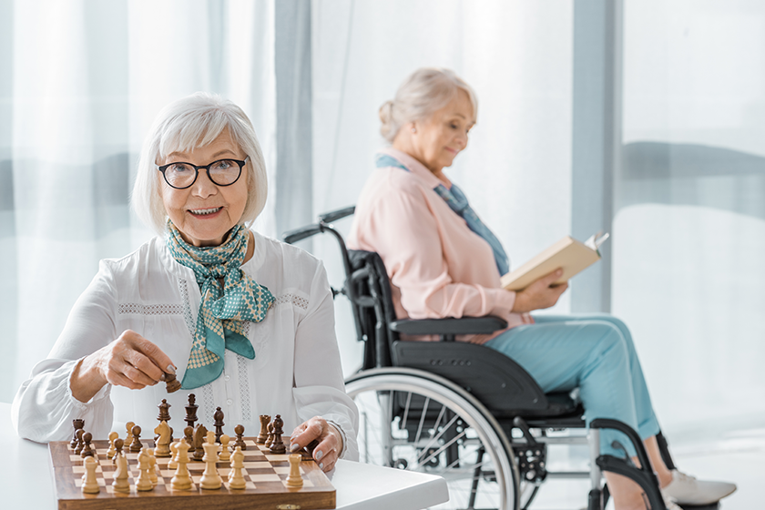 Senior woman playing chess at table while another siting wheelchair reading Senior woman playing chess at table while another siting wheelchair reading