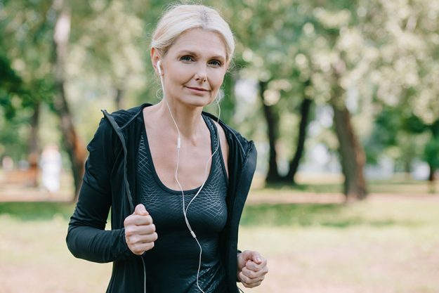 beautiful mature sportswoman smiling camera while running park listening music