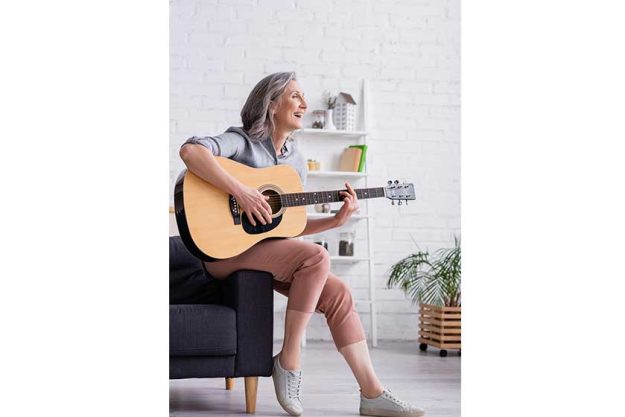 cheerful mature woman grey hair playing acoustic guitar while sitting