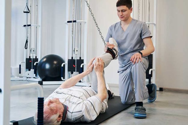 Doctor pulling right leg of pensioner with attached resistance band