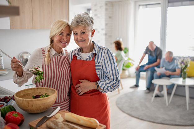 Group of senior friends having party indoors cooking and looking at camera